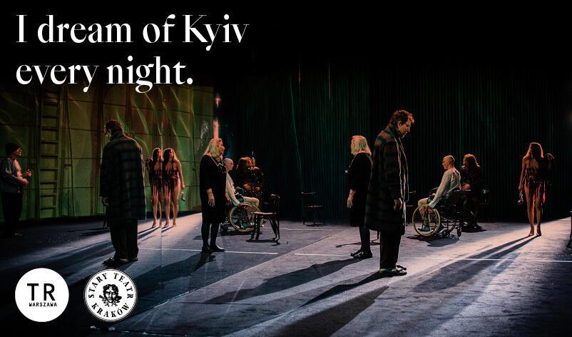 People are standing on the stage. Some are on wheelchairs. They look in different directions. Sign says: I dream of Kyiv every night. This is an image from 3Siostry (3Strs). Dir. by Luk Perceval. 