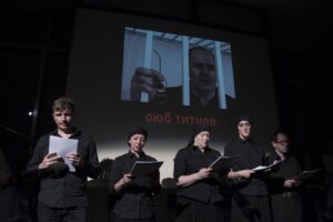 Five actors, all in black, stand in front of a projected image of a man who is behind bars. The name Oyub Titiev appears in red beneath his image.