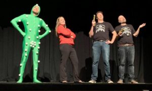 Against the black backdrop, four actors of the 2019 fringe San-Diego production of Space Force! stand on a stage. One is wearing a light green attire designed for filming actors for the CGI, others are wearing casual informal clothing.