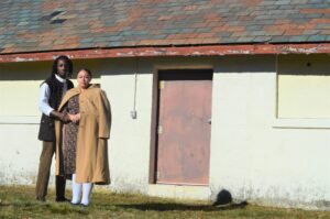 Outdoors photograph of two black actors, Brionna Bonet and Stephen Sampson, standing in from of a barn. The actors are wearing modernized historical clothing as their costumes for The Toby Gilmore Story.