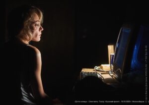 Photo of a caucasian actress seating in the dim light, profile facing a desk with a computer monitor on it.