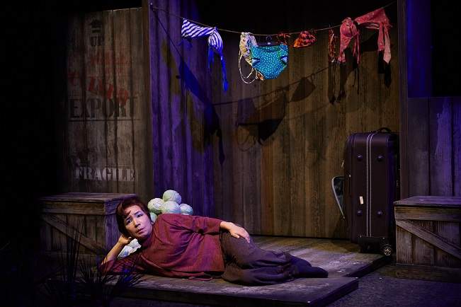 A woman lying on the floor in a bare room, with a suitcase and laundry clothesline in the background.