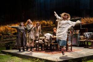 An image of a woman dancing while other women are clapping. There is a family table and the scenography suggests rural setting.