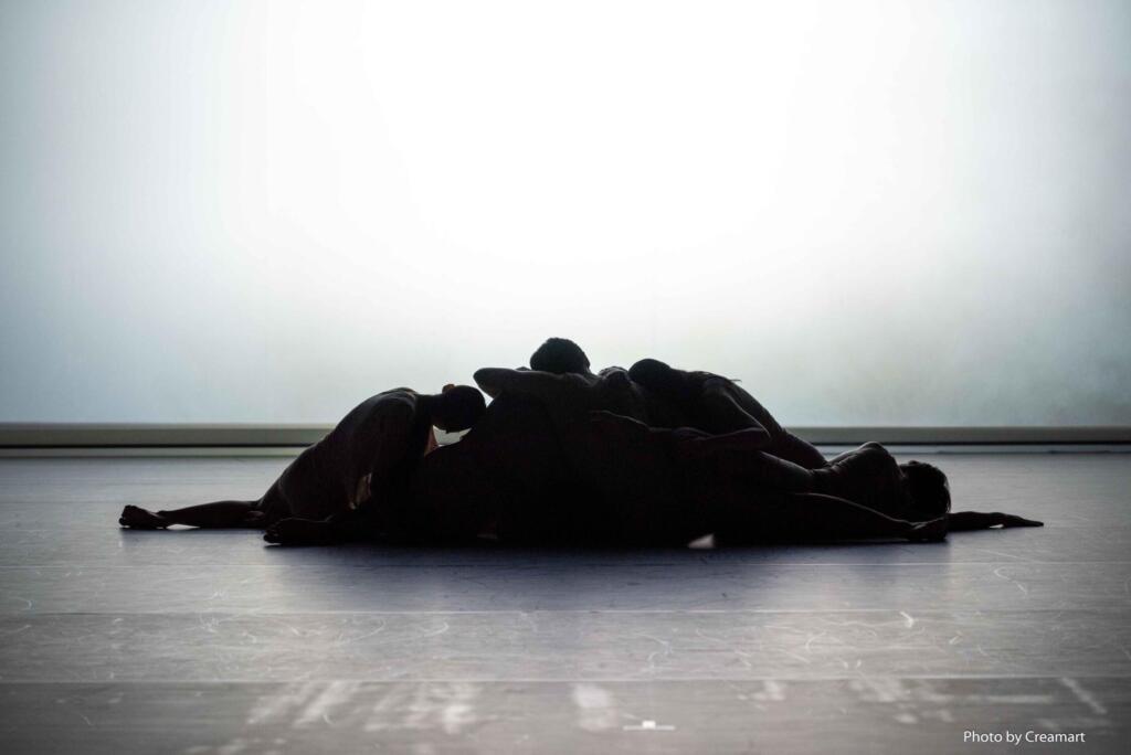 A group of bodies huddling together on the floor of a bare stage.