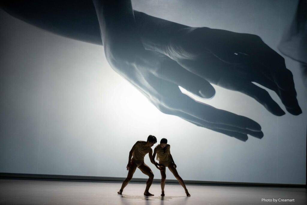 Two dancers are on a bare stage. Their maginfied hands are projected onto the back wall.