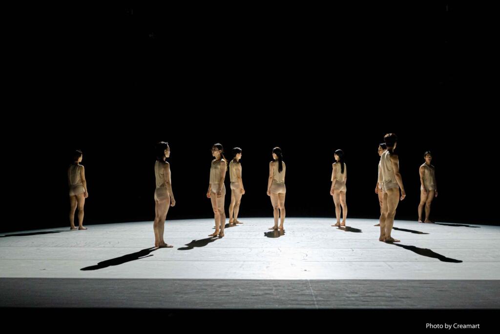 A group of dancers are standing still on a bare stage.