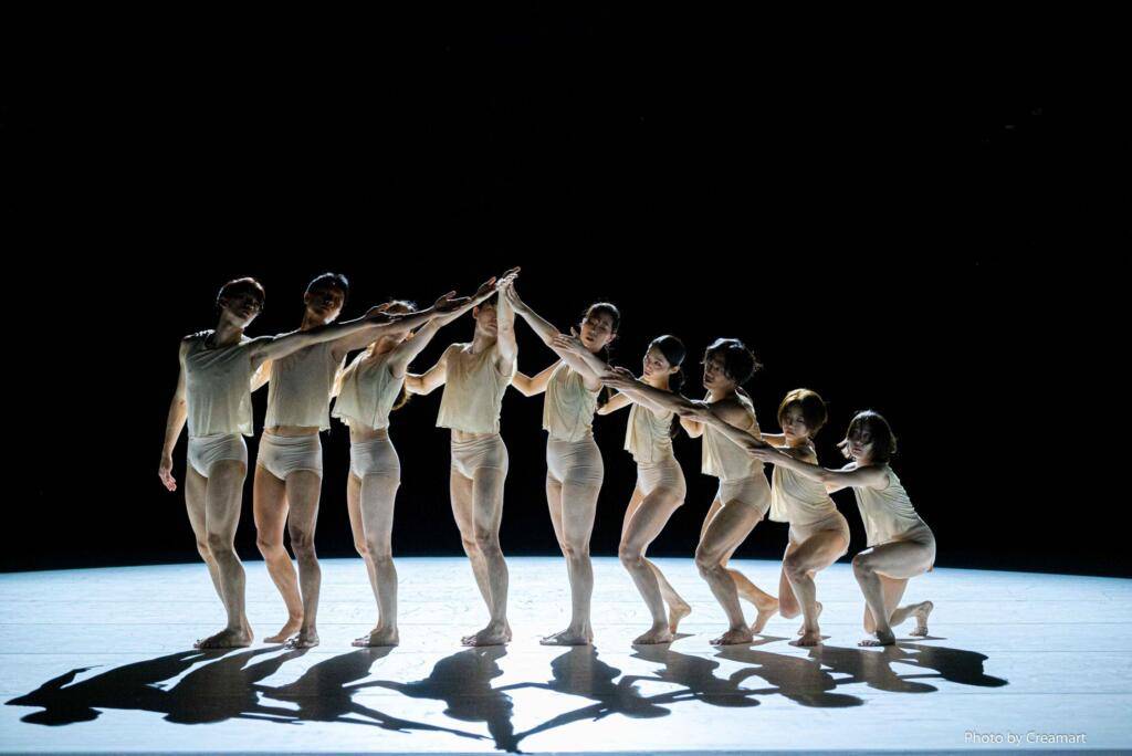 A group of dancers are on a bare stage. Their arms are linked together, creating a curved line.