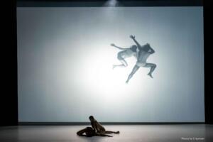 Two dancers are on the ground on a bare stage. Their image is projected onto the back wall.